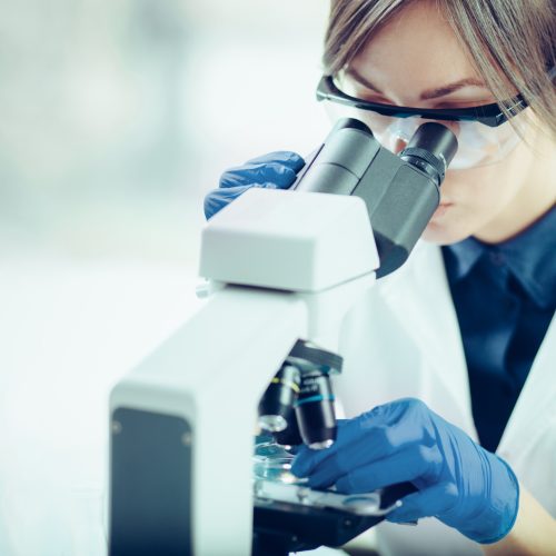 A scientist wearing safety goggles and blue gloves closely examines a sample through a microscope in a laboratory setting.