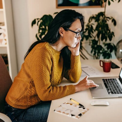 A woman in a yellow sweater sits at a desk, looking intently at a laptop screen. She is resting her head on one hand, with a notebook, mug, and mouse on the desk in a cozy home office setting.