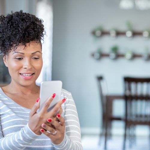 A woman with curly hair and red nails, wearing a gray and white striped shirt, smiles while looking at her smartphone in a bright kitchen or dining area with plants on the wall in the background.