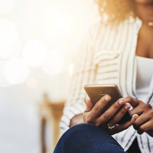 A person wearing a striped shirt sits with legs crossed and uses a smartphone. The background is bright and softly blurred, creating a warm, casual atmosphere.