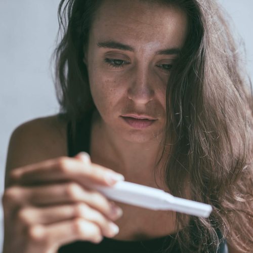A woman with long brown hair looks intently at a pregnancy test she is holding, her expression appearing serious and contemplative.