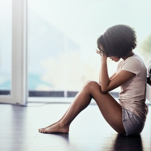 A woman sits on the floor next to a bed with her knees drawn up and her head in her hands, appearing upset or deep in thought. Sunlight filters through large windows in the background.