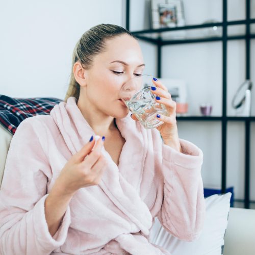 A woman in a pink bathrobe sits on a couch, holding a pill in one hand and drinking a glass of water. She appears relaxed in a cozy home setting with shelves and framed photos in the background.