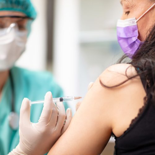 A healthcare worker wearing a mask, face shield, and gloves administers a vaccine injection into the upper arm of a woman who is also wearing a mask.