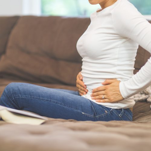 A woman sits on a couch holding her stomach with both hands, possibly experiencing abdominal discomfort. An open book lies next to her on the brown sofa.
