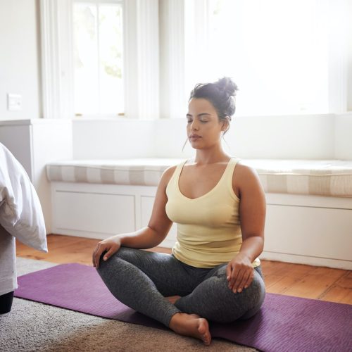 A woman sits cross-legged on a purple yoga mat in a bright room, eyes closed and hands resting on her knees, appearing calm and meditative. Sunlight streams through large windows behind her.