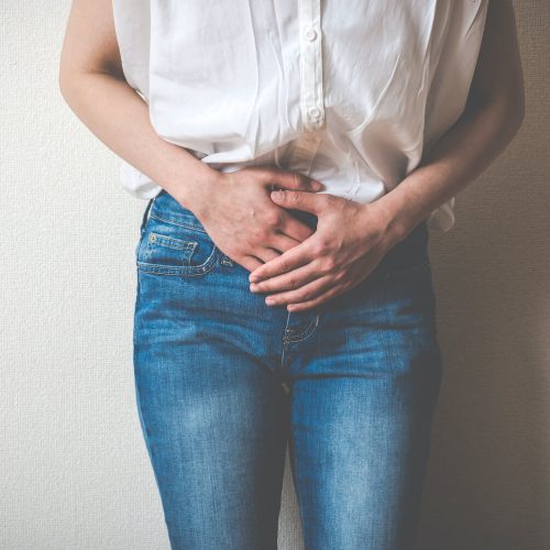 A person wearing a white shirt and blue jeans stands against a wall, holding their lower abdomen with both hands, suggesting discomfort or abdominal pain.