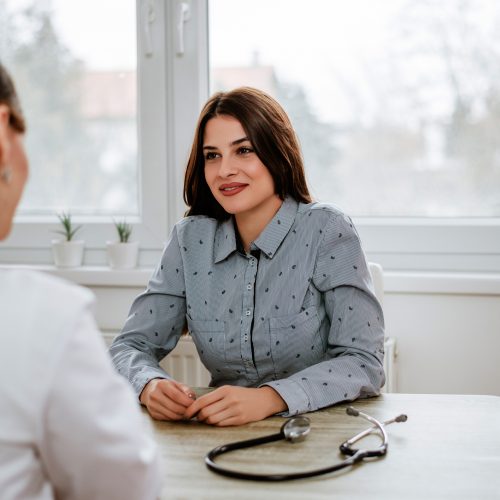 A woman in a blue shirt sits at a desk, smiling and talking with a doctor whose back is turned. A stethoscope lies on the desk in a bright, modern office.