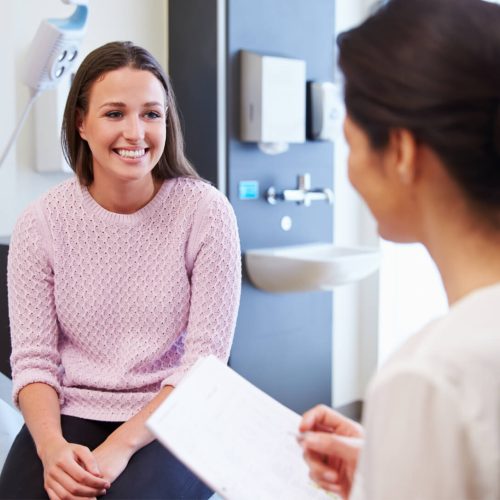 A young woman in a pink sweater smiles while sitting on an exam table, talking to a healthcare professional who is holding a clipboard in a medical office.