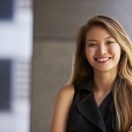 A young woman with long, light brown hair smiles confidently, standing with arms crossed in a sleeveless black top, in a modern indoor setting with blurred background.