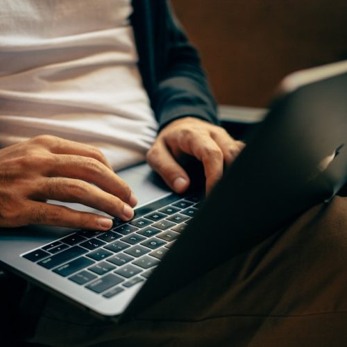 Close-up of a person typing on a laptop. They are wearing a white shirt, dark jacket, and brown pants, and are seated with the laptop resting on their lap. The focus is on their hands and the keyboard.