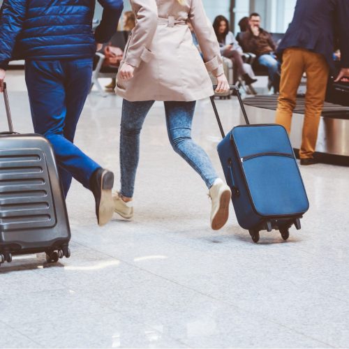 Two people walk quickly with rolling suitcases through an airport baggage claim area. Other travelers are sitting and standing in the background, and luggage carousels are visible.