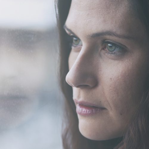 A woman with long brown hair looks pensively out of a window, her face partially reflected in the glass. The mood is contemplative and somber, with soft natural light illuminating her features.