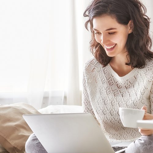 A woman sits on a bed, smiling while using a laptop. She is holding a white mug, surrounded by pillows and flowers in a bright, cozy room with natural light.