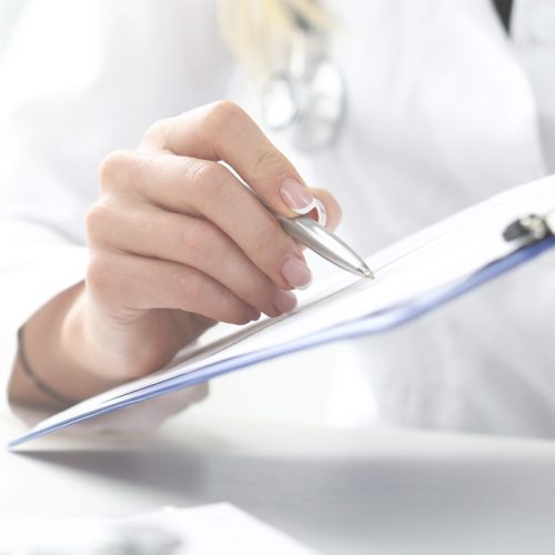 A doctor in a white coat holds a clipboard and pen, discussing medical information with a patient whose hands are folded on the table.
