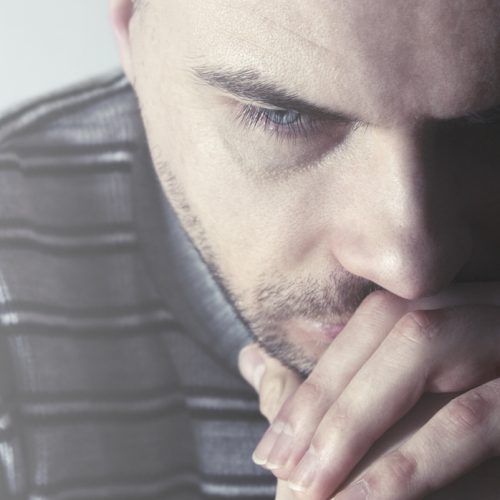 A man in a striped sweater sits with his hands pressed to his mouth, looking downward with a serious and contemplative expression.