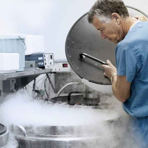 A man in blue scrubs lifts the lid of a large cryogenic storage tank emitting vapor in a laboratory setting with scientific equipment and containers visible.