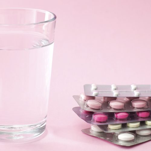 A clear glass of water sits next to four blister packs of various pink and white pills on a light pink background, symbolizing the options and support available, such as egg freezing insurance, for those considering their reproductive health.