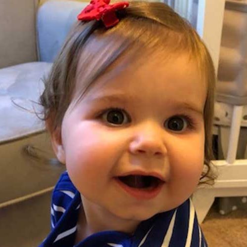 A smiling baby with brown hair, wearing a blue and white striped shirt and a red bow hair clip, sits in front of a white crib and a beige chair.