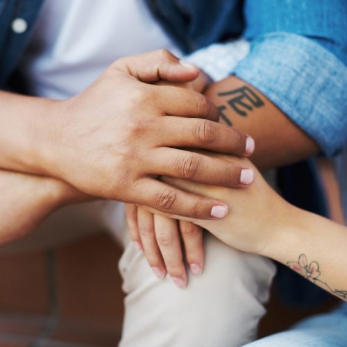 Close-up of two people sitting closely, gently holding hands in a comforting gesture. One person has a tattoo on their forearm and the other has a floral tattoo on their wrist. Both are wearing denim shirts.