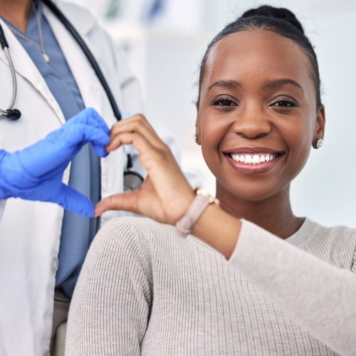 A smiling woman and a doctor wearing a white coat and blue gloves form a heart shape with their hands, symbolizing care and support, in a bright medical setting.