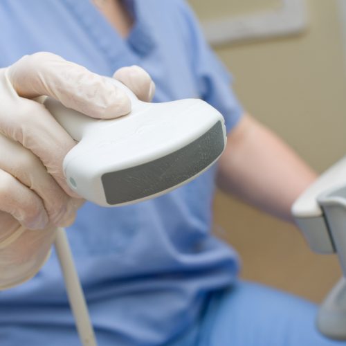 A close-up of a healthcare worker wearing gloves and holding an ultrasound probe, with part of the ultrasound machine visible in the background.