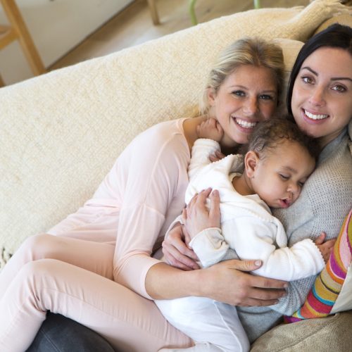 Two women sit closely together on a beige couch, smiling at the camera, while holding a sleeping baby in their arms. The scene appears warm and affectionate, with soft natural light.