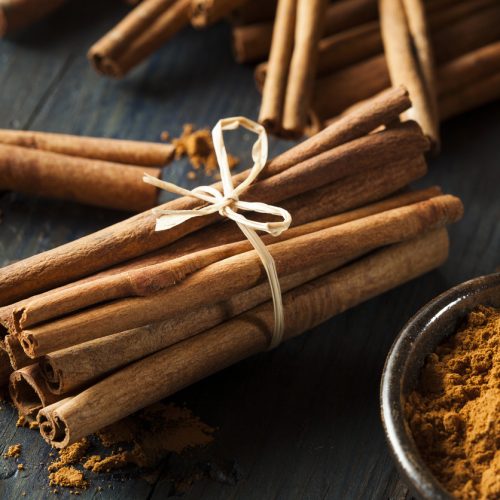 A bundle of cinnamon sticks tied with twine sits on a wooden surface next to a bowl of ground cinnamon, with more cinnamon sticks and scattered powder in the background.