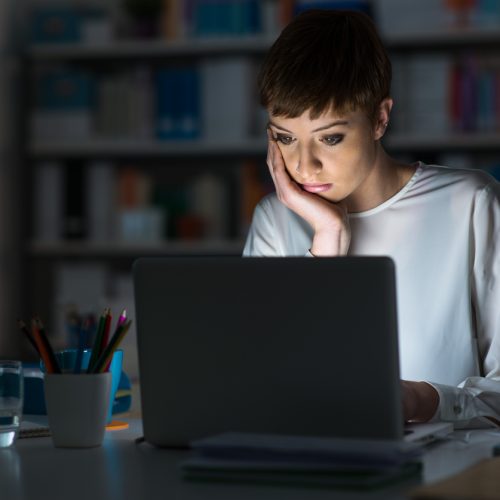 A woman sits at a desk late at night, looking intently at her laptop screen with her hand on her face. The room is dimly lit, and there are office supplies and a glass of water on the desk.