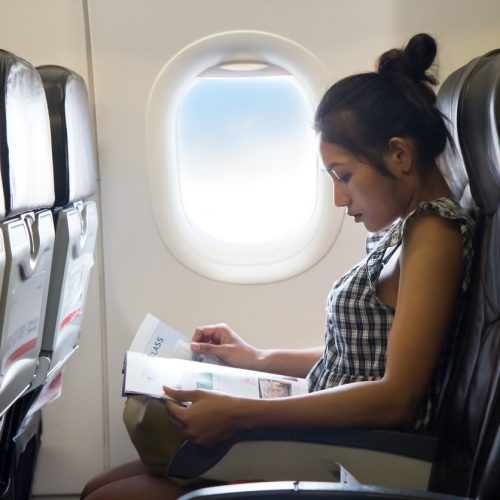 A woman sits in an airplane seat by the window, reading a magazine. Sunlight streams through the window beside her, and the cabin appears calm and quiet.