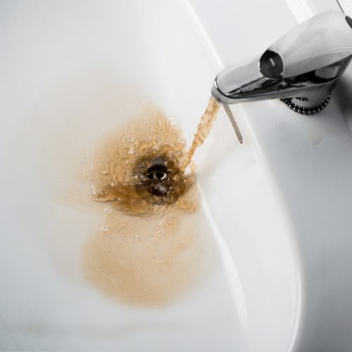 Brown, dirty water flows from a faucet into a white sink, swirling around the drain, indicating contaminated or rusty tap water.