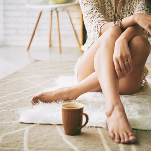 A woman sits on a white fluffy rug with her legs crossed, wearing a knit robe. Her face is not visible. A brown mug filled with a hot drink is placed on the rug in front of her.