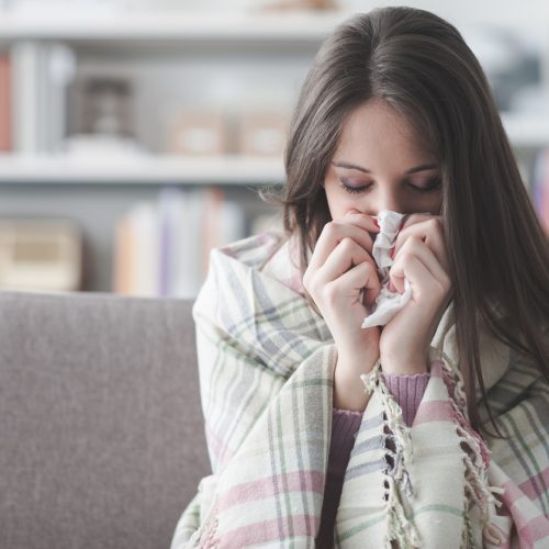 A young woman wrapped in a plaid blanket sits on a couch, holding a tissue to her nose as if she is sneezing or has a cold. The background shows a softly focused living room.