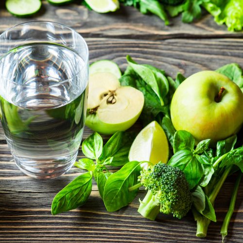 A glass of water sits on a wooden table surrounded by green apples, spinach leaves, broccoli, lime wedges, basil, and leafy greens. The fresh produce creates a healthy and vibrant arrangement.
