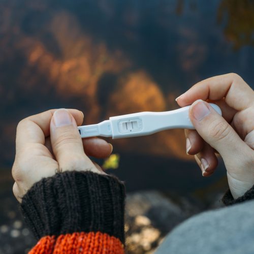 Close-up of hands holding a pregnancy test with one line visible, indicating a negative result. The person is wearing a dark sweater with an orange sleeve, and there is a blurred outdoor background.