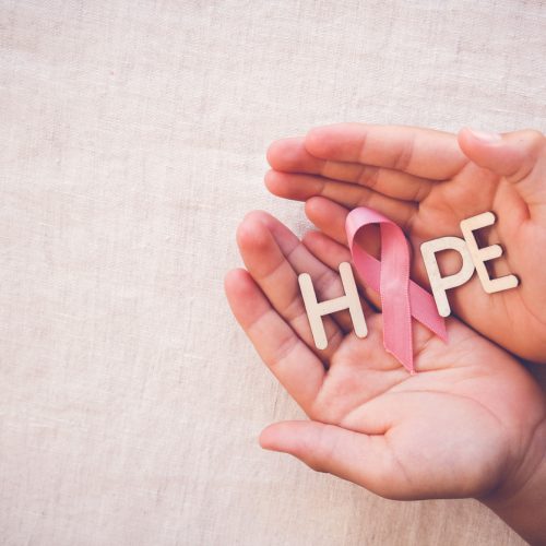 Two hands gently hold wooden letters spelling “HOPE” with a pink ribbon, symbolizing breast cancer awareness, on a light fabric background.