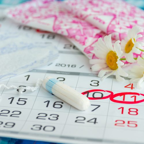 A calendar with several dates circled in red, a white tampon, sanitary pads in pink packaging, and white daisies laid on top, representing menstruation and period tracking.