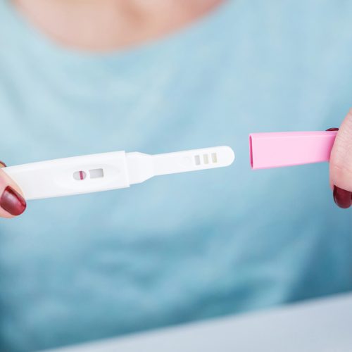 A person holding a pregnancy test with two visible lines, indicating a positive result. The person’s fingernails are painted red and they are wearing a light blue shirt.
