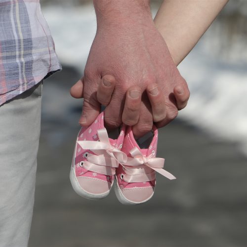 A couple holds hands while grasping a pair of small pink baby shoes, symbolizing an upcoming addition to the family. The man wears plaid, and the woman wears a floral dress.