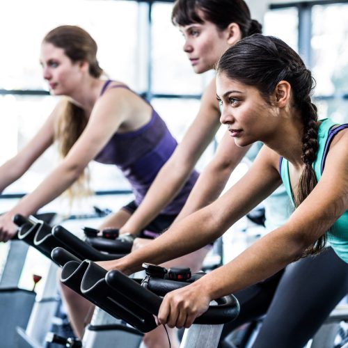 Three women in athletic wear ride stationary bikes in a bright gym, focused and determined, exercising together in a row.
