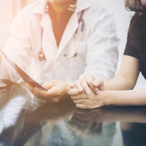 A doctor in a white coat discusses medical results with a patient, holding a paper while the patient clasps their hands together on a table, suggesting a comforting or supportive conversation.