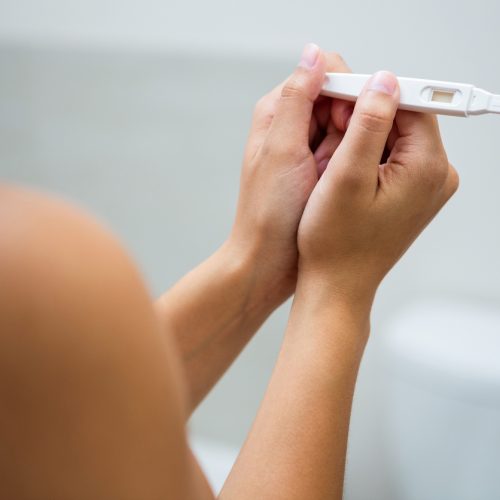 A person is holding a pregnancy test with both hands, looking at the result. The background is out of focus, showing a bathroom setting.