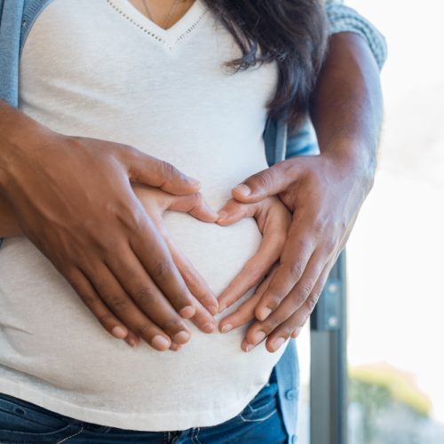 Close-up of a pregnant woman’s belly with two pairs of hands gently forming a heart shape over it, symbolizing love, support, and anticipation for the baby.