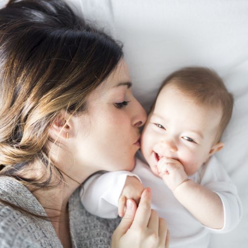 A woman lies on a bed, gently kissing a smiling baby on the cheek. The baby is wearing a white outfit and looks happy, holding the womans hand. Both are resting on white bedding.
