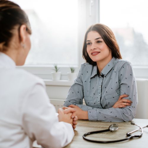 A woman in a light blue shirt sits across from a doctor in a white coat, smiling and talking in a bright office. A stethoscope lies on the desk between them.