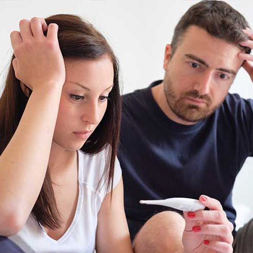 A worried-looking couple sits close together, both touching their heads with concerned expressions, as the woman holds and looks at a pregnancy test.