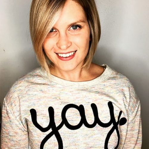 A smiling woman with straight blonde hair wears a light-colored sweater with the word yay. written in large black script across the front, standing against a plain background.
