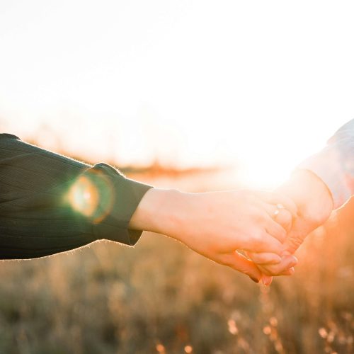 Two people holding hands outdoors at sunset, with warm sunlight creating a soft, glowing effect in the background.