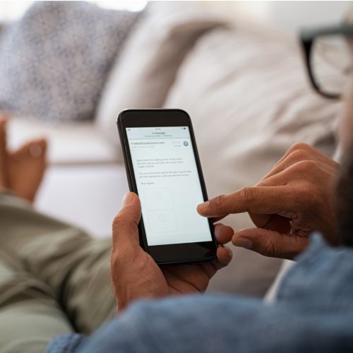 A person sitting on a couch uses a smartphone, browsing a website or app. The individual holds the phone in one hand and interacts with the screen using the other, with legs crossed and a relaxed posture.