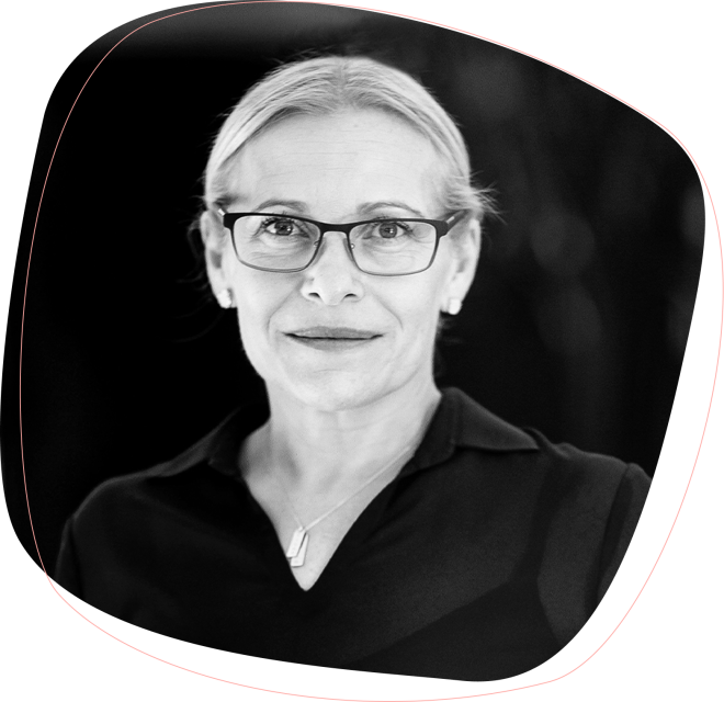 A woman with light hair pulled back, wearing glasses, a dark collared shirt, and a necklace, looks at the camera with a neutral expression. The image is black and white with a rounded border.